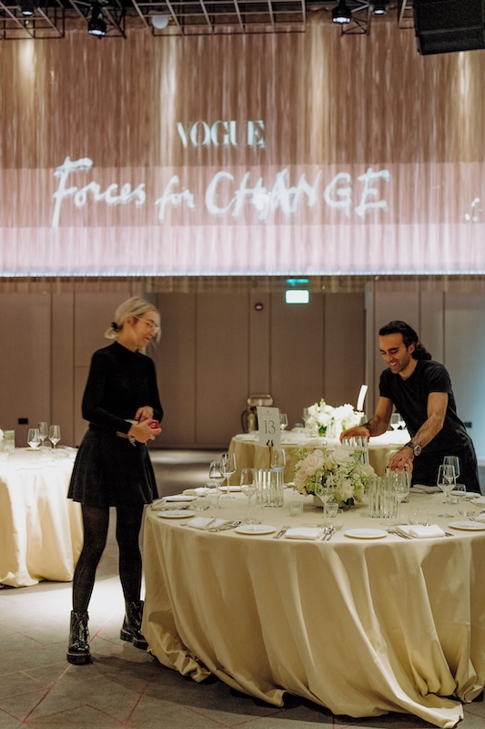 Two people prepare a round dining table adorned with a white tablecloth and wedding flowers expertly arranged by a luxury London florist. Wine glasses, silverware, and dishware are set, with a table number 13 stand in the middle. Behind them, a large screen displays "Vogue Forces for Change." The room is softly lit.