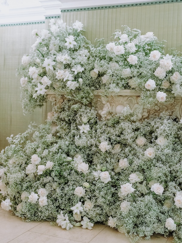 A lavish floral display featuring an abundance of white roses, lilies, and baby’s breath cascading from a large, ornate, stone planter. Curated by a luxury florist in London, the arrangement creates a lush, overflowing effect against a pale green wall with white molding. The overall aesthetic is elegant and luxurious.