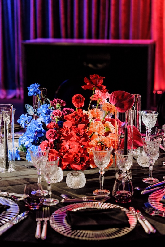 A lavish table setting features an elegant arrangement of red, blue, and orange wedding flowers as the centerpiece. The table is adorned with crystal glassware, silver cutlery, and intricately patterned plates with black napkins. The backdrop showcases a curtain bathed in red and blue lighting.