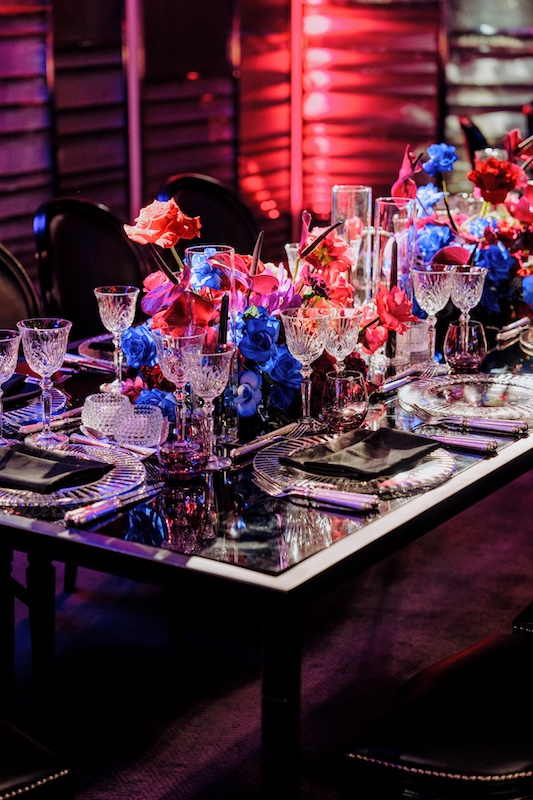 A beautifully set dining table with an elegant and vibrant arrangement. The table features crystal glassware, black napkins on clear plates, and a stunning floral centerpiece with red, pink, and blue flowers from a luxury London florist. The ambient lighting casts a warm glow, enhancing the sophisticated atmosphere.