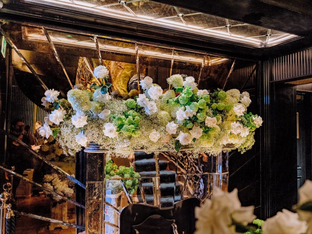 A lavish floral arrangement of white and green flowers by a luxury florist sits atop a black marble fireplace in an opulent room. The mirror behind the fireplace reflects more flowers, adding depth to the scene. The room features elegant decor with warm lighting, creating a luxurious and sophisticated atmosphere.