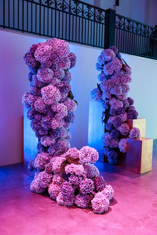 A vibrant art installation by a luxury London florist featuring three tall, abstract sculptures composed of clusters of purple flowers. They are placed on metallic pedestals in front of a white wall and under blue and purple lighting. A black metal railing is visible in the background above the setup.