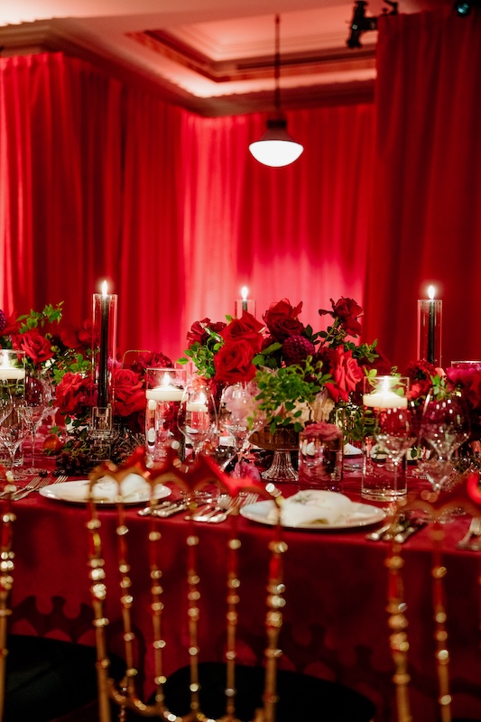 A lavish banquet setup features a long table adorned with elegant event flowers in red and green, numerous candles in glass holders, and fine china. Gold chiavari chairs surround the table, and the room is illuminated by red draped curtains and a hanging light fixture provided by a luxury London florist.