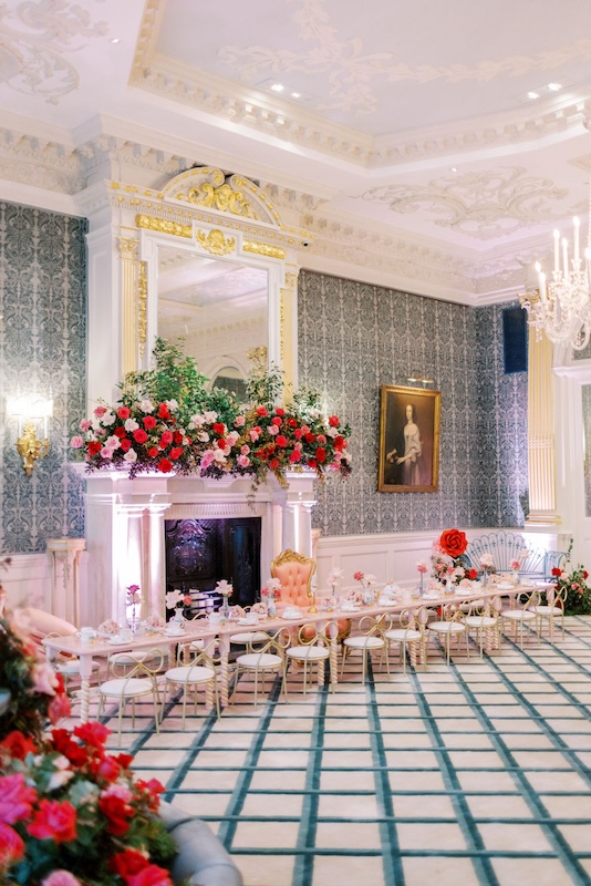 An elegantly decorated room features a long table set for a formal tea party. The table is adorned with teacups, saucers, and floral arrangements from a luxury florist. A grand fireplace with a large mirror and lush flower display stands behind the table. The room has ornate wallpaper, and a chandelier hangs from the ceiling.