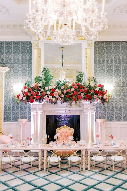 A lavish, elegant room features a large marble fireplace adorned with a lush floral arrangement by a luxury London florist. The room has ornate wallpaper, chandeliers, and a long dining table set with fine china. Gold-framed chairs line the table, and a plush, golden chair sits at the head.