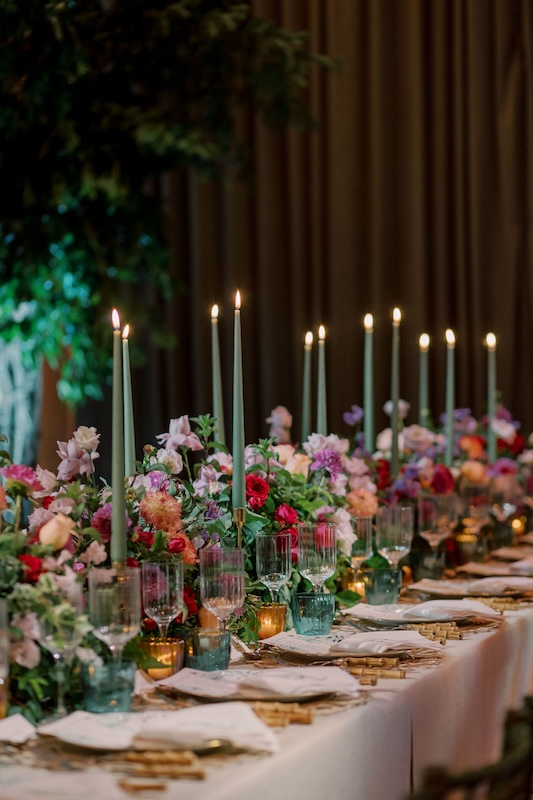 Elegant table setting with tall, lit green candles in candleholders lined down the center of the table. The table is adorned with vibrant event flowers featuring reds, pinks, purples, and whites. Crystal glassware and folded napkins complete the sophisticated look.