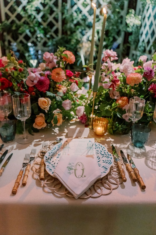 A dining table decorated with vibrant floral arrangements from a luxury London florist, ornate gold candle holders with lit green candles, elegant glassware, and cutlery with bamboo-style handles. The centerpiece includes a folded napkin with a green monogram "O" placed on a decorative plate with a leaf pattern.