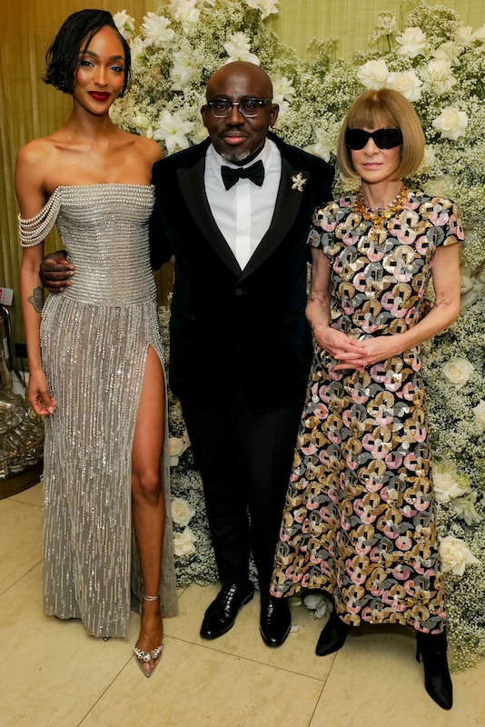 Three people are posing for a photo in front of a stunning floral backdrop curated by a luxury London florist. On the left, a woman in a silver off-shoulder gown with thigh-high slit; in the middle, a man in a black tuxedo wearing glasses and a bow tie; on the right, a woman with bob haircut wearing shades and floral dress.