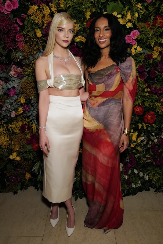 Two women stand in front of a lush floral wall. The woman on the left has long blonde hair, a metallic silver bandeau top, and a cream skirt paired with white heels. The woman on the right, framed by luxurious London flowers, has long dark hair and wears a colorful one-shoulder dress with shades of red, purple, and beige.