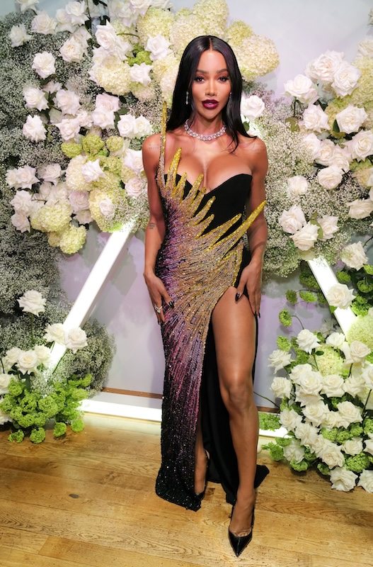 A woman in a sleek black and gold dress poses on a wooden floor in front of a floral wall arrangement by a luxury London florist, featuring white and light green flowers. The dress has a high slit, revealing her left leg. She wears a diamond necklace and black high heels, with her long dark hair styled straight.