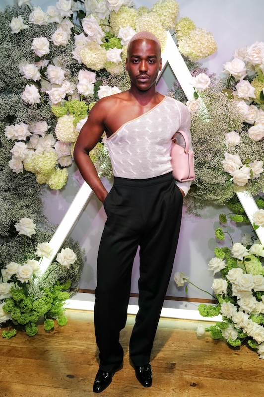 A person stands in front of a stunning floral backdrop created by a luxury florist, featuring white and green flowers. They are wearing a one-shoulder, semi-sheer white top paired with high-waisted black pants and shiny black shoes. Holding a small, light pink handbag, they exude confidence and poise.