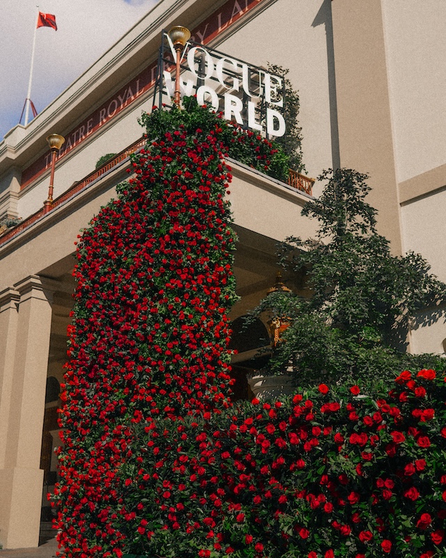 A building with a "Vogue World" sign on top, featuring an extensive display of lush red roses cascading down the façade like a London florist's masterpiece. The structure has classic architectural elements, including columns and a curved arch. A flag is visible atop the building on the left side.