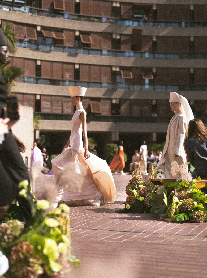 A fashion show takes place outdoors on a sunlit brick pathway. Models showcase avant-garde outfits, including long dresses and tall, structured headpieces. The backdrop features a curved, modern building with open windows. Vibrant London flowers decorate the path's edges, adding to the event’s charm.