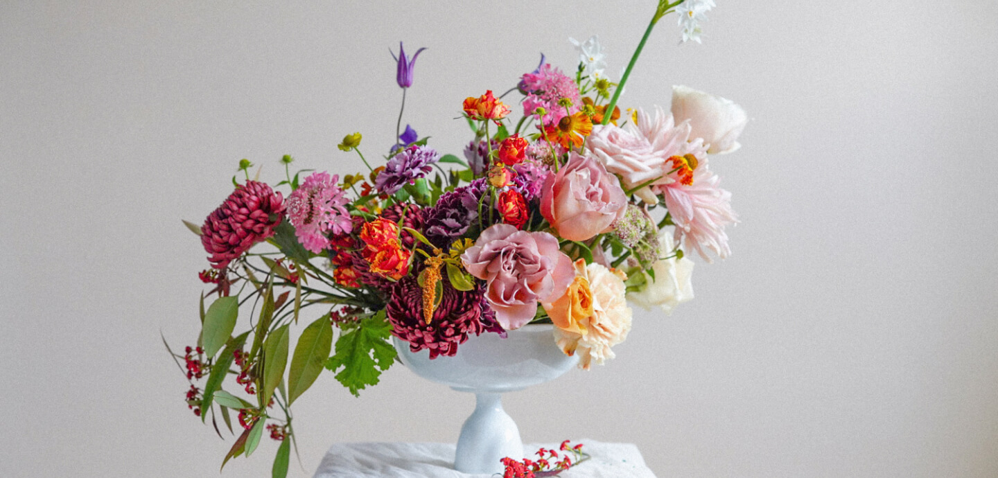 A white pedestal vase from a luxury London florist holds an assortment of colorful flowers, including pink and peach roses, red zinnias, and purple clematis. Green leaves and small red berries cascade out of the arrangement, which sits on a white table against a simple light gray background.