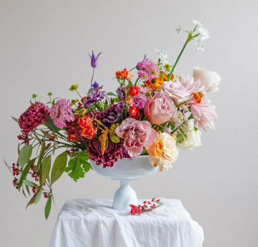 A white ceramic vase brimming with a diverse arrangement of London flowers sits atop a draped white cloth on a table. The bouquet includes pink and peach roses, purple flowers, and red berries, creating a vibrant and elegant display against a muted background.