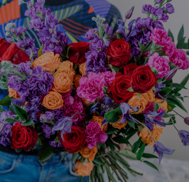 A person in a colorful, patterned shirt holds a large bouquet of vibrant flowers, including red roses, purple stock, pink carnations, orange spray roses, and purple lisianthus. The mix of blooms creates a rich, colorful arrangement against a neutral background.