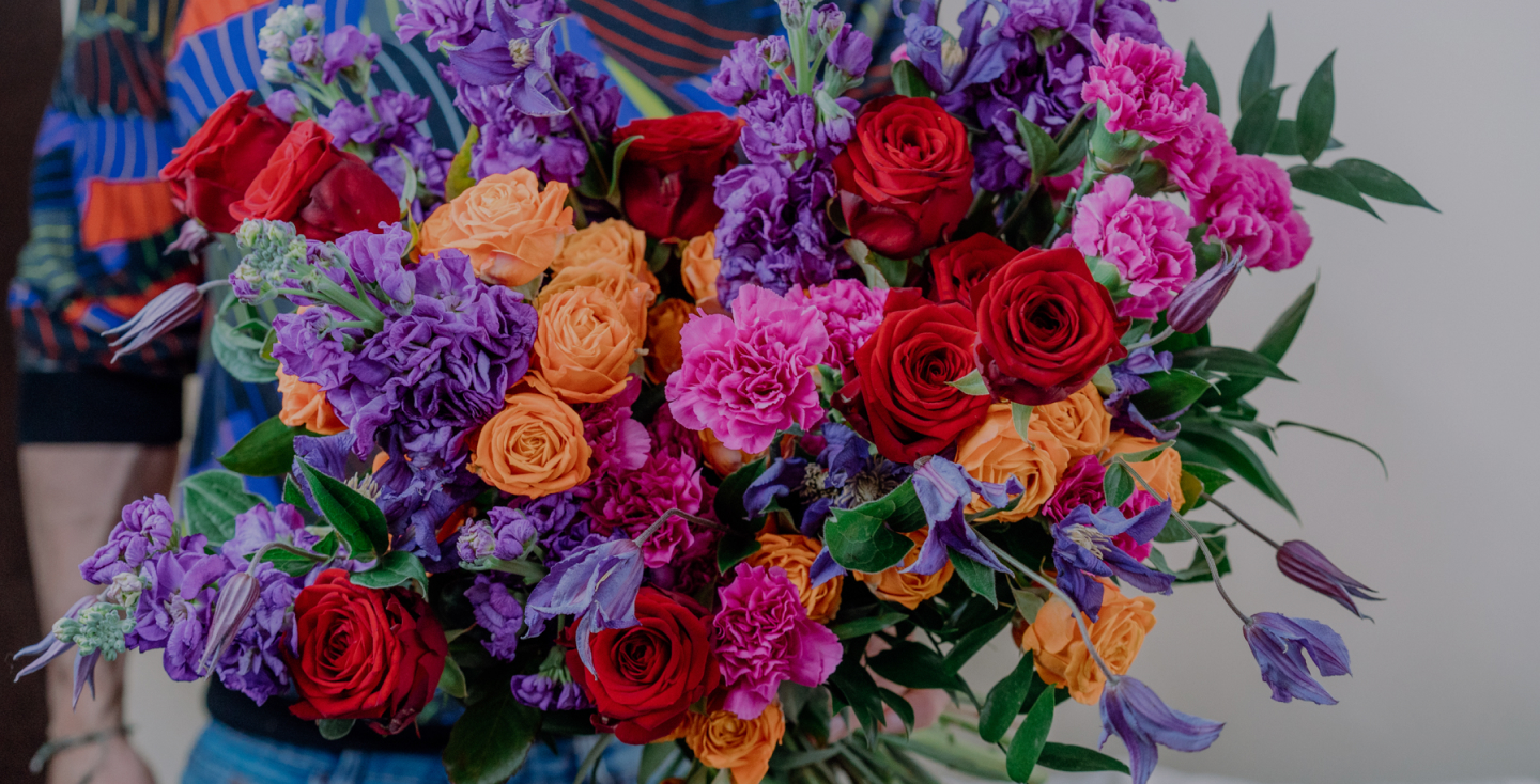 A vibrant bouquet featuring red roses, orange roses, pink carnations, and purple flowers. The mix of colors creates a striking contrast, accented by lush green leaves. A person in a colorful outfit holds the bouquet against a neutral background.