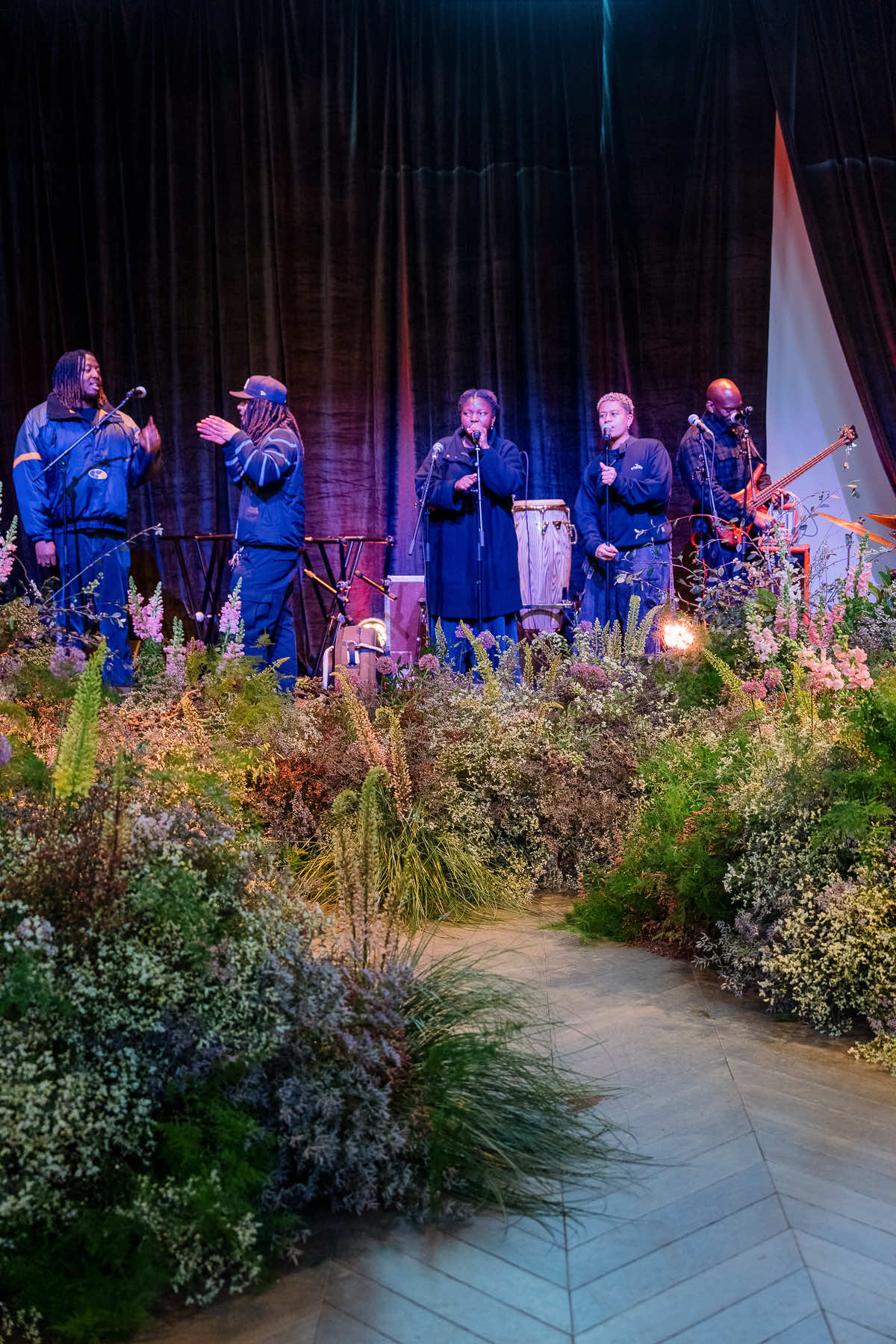 A musical group in matching blue outfits performs on stage with a backdrop of dark curtains, surrounded by lush greenery and corporate event flowers. Five members sing into microphones, and one plays a guitar. The setting appears vibrant and lively, emphasizing their energetic performance.