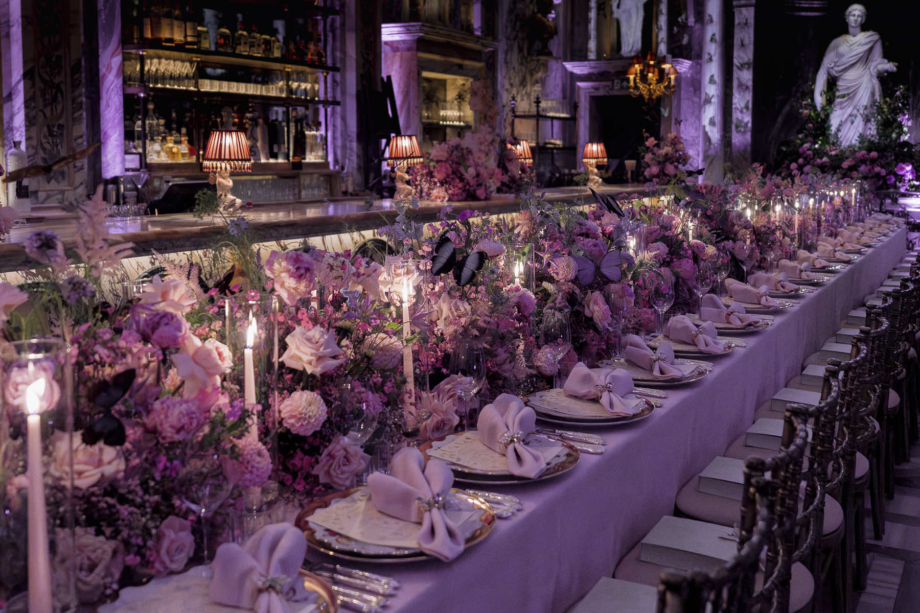 An elegantly set long dining table with intricate floral arrangements in shades of purple and pink, masterfully arranged by a corporate florist. The table features white napkins, gold-rimmed plates, and lit candles. Behind, there are shelves lined with bottles, a statue, and purple ambient lighting, creating a luxurious atmosphere. Photo taken in Bacchanalia, London.