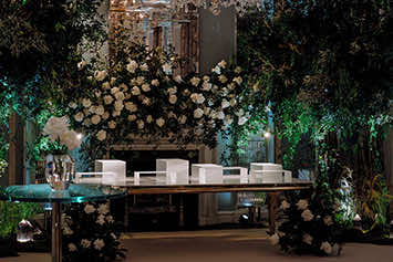 A venue adorned with lush greenery and abundant white flowers, expertly arranged by an event florist. A transparent glass table is at the center, with white chairs arranged behind it. The backdrop features cascading flowers, and the ambient lighting casts a warm and inviting glow over the elegant setting.