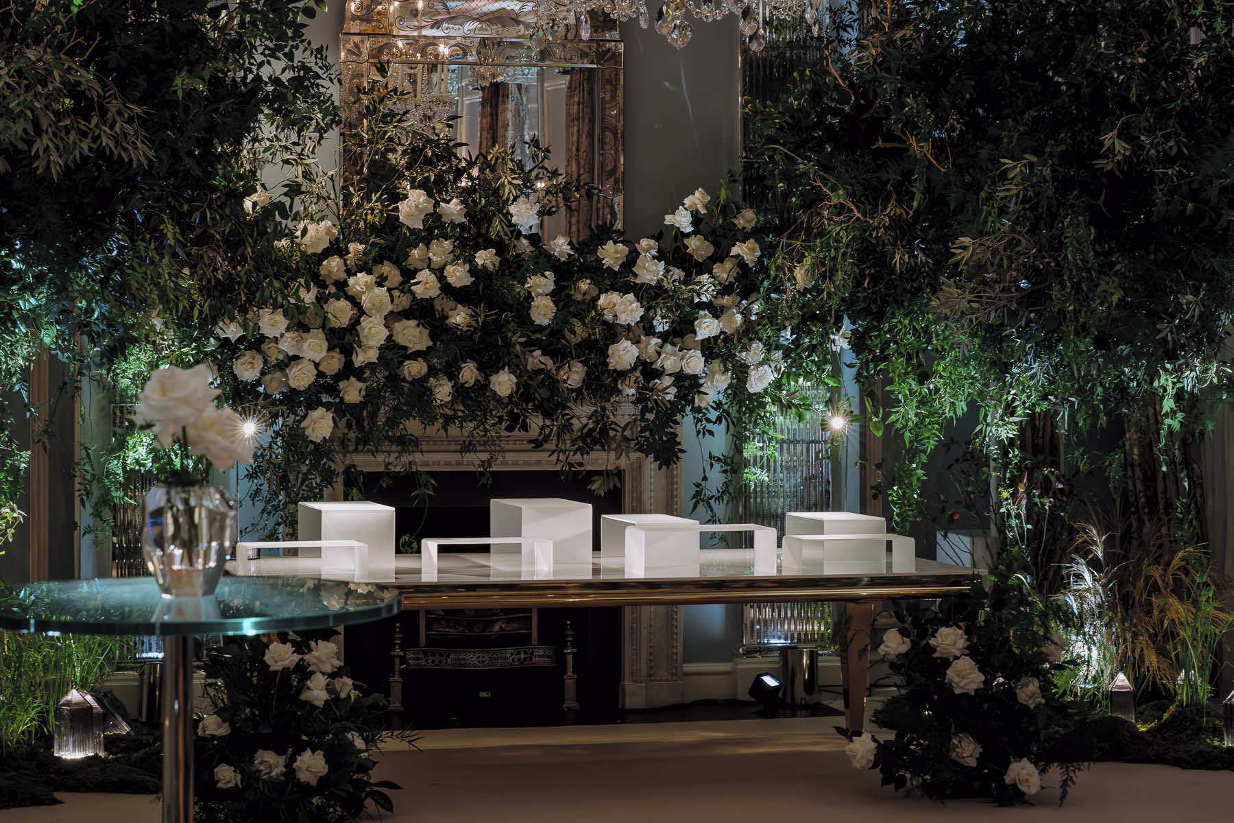 An elegant indoor setting features a lavish floral arrangement with white flowers and lush greenery surrounding a fireplace, crafted by an event florist. A reflective table with a single white flower in a vase is in the foreground, while four stylish white chairs are positioned on a platform in the background.