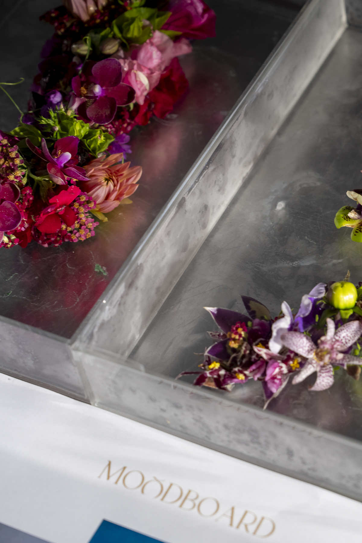 The image shows two floral arrangements on a gray, textured surface with transparent dividers. One arrangement features pink, red, and purple flowers, while the other highlights purple and green blooms. "MOODBOARD" is written on a visible white card at the bottom—a testament to a skilled event florist.