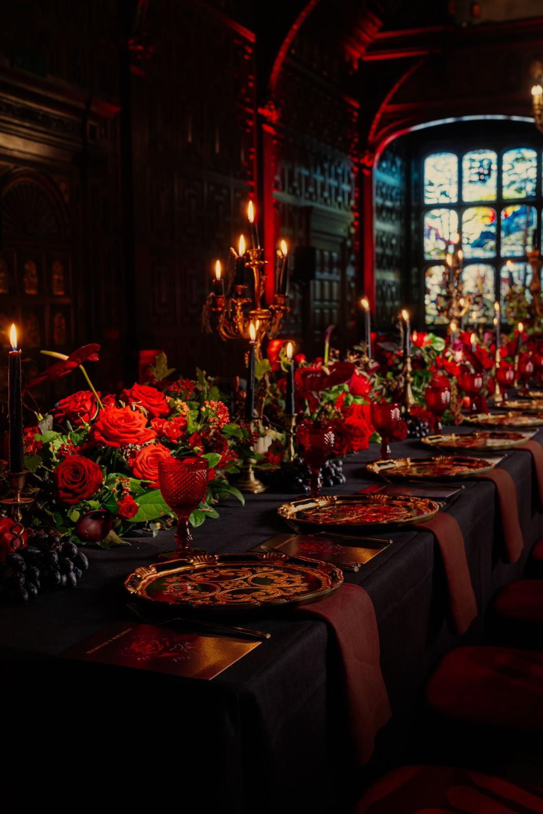 A richly decorated dining table is set with red roses, greenery from the event florist, gold-rimmed plates, and black candles in ornate holders. The scene is illuminated by warm, dim lighting with red accents highlighting the intricate wood-paneled walls and stained glass windows in the background.