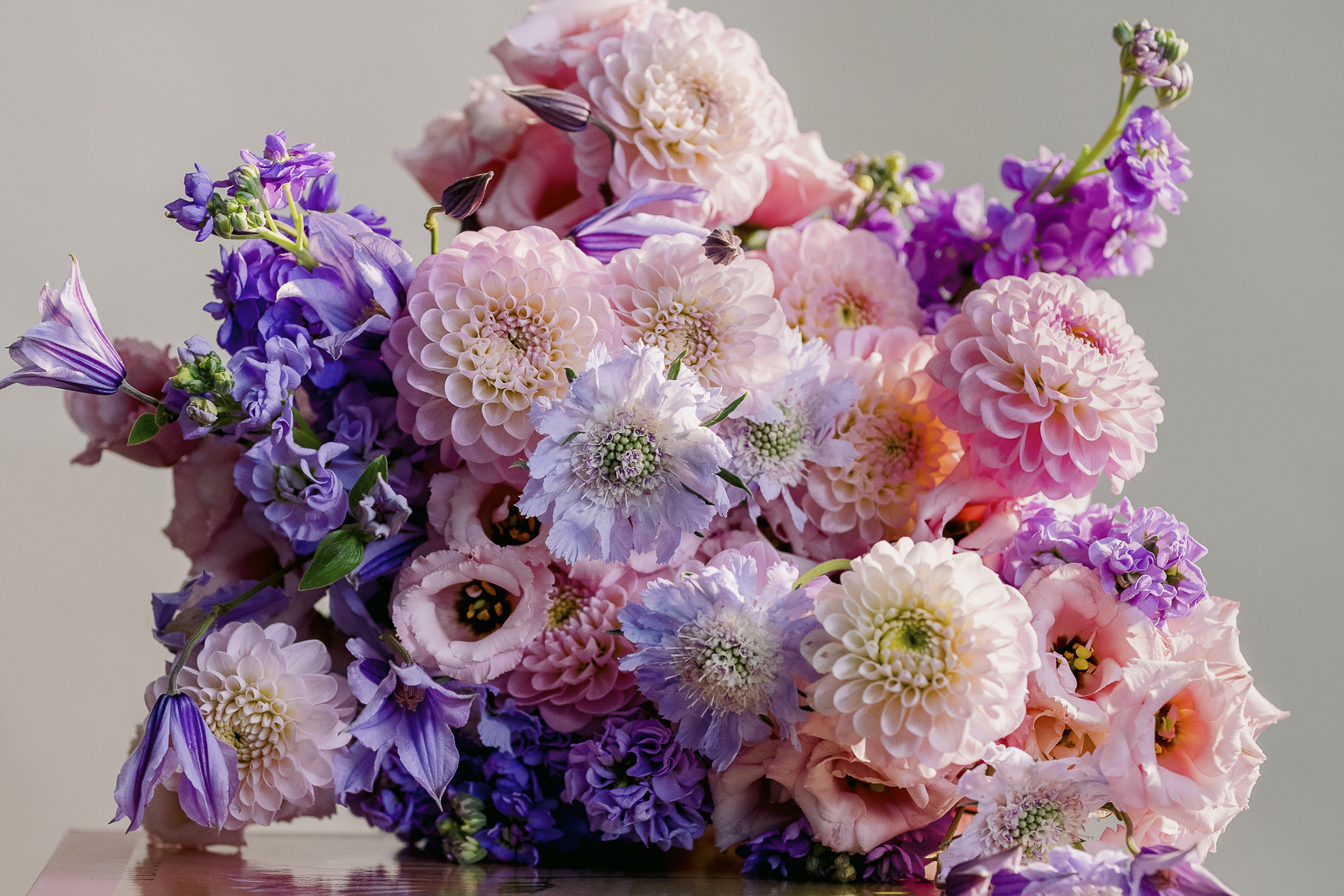 A lush bouquet of mixed flowers featuring pink and lavender blooms crafted by a luxury florist. The arrangement includes dahlias, lisianthus, stock flowers, and clematis, with varied petal textures and shades. Displayed on a reflective surface, the vibrant colors shine against a neutral background.